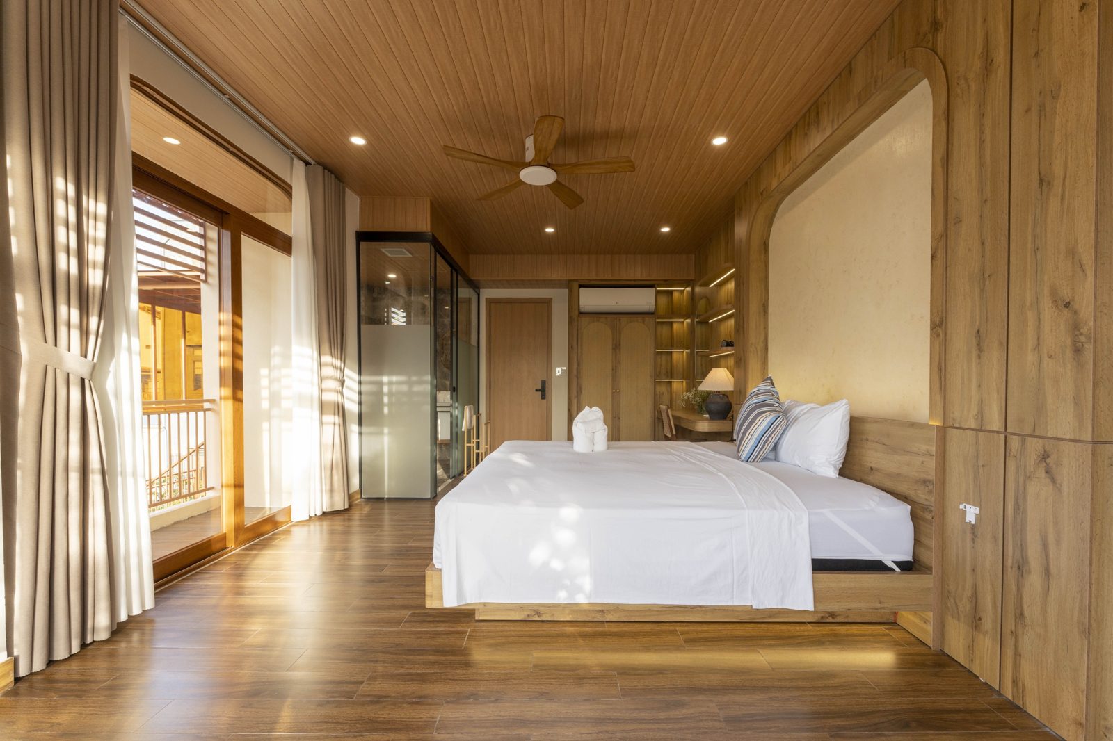 Long bedroom with balcony doors, wooden fan and glass-walled bathroom
