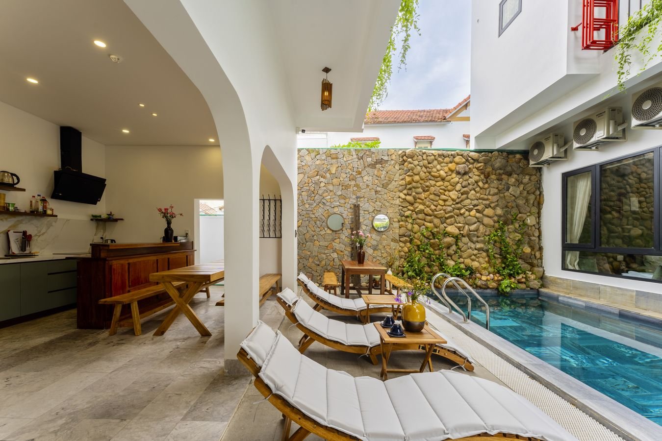Pool beside a stone wall with white loungers and an open-air kitchen